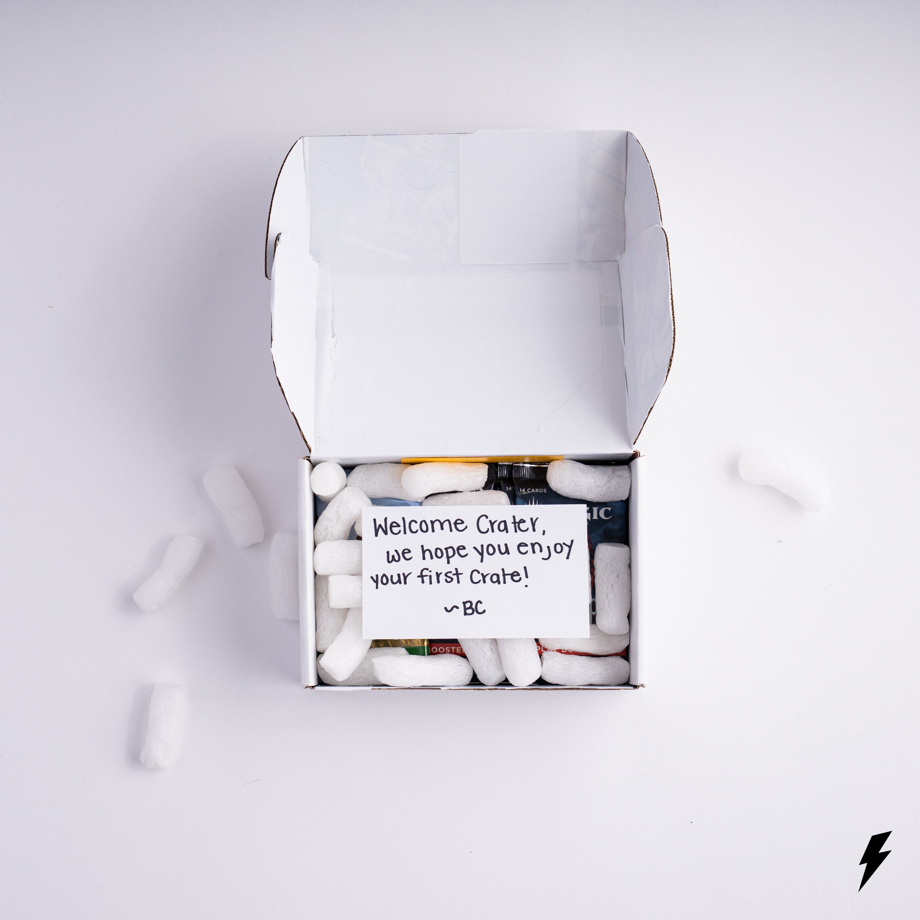 Open white Booster Crate box filled with packing peanuts and trading card packs, featuring a handwritten note that reads, “Welcome Crater, we hope you enjoy your first Crate! ~BC” placed on top inside the box.